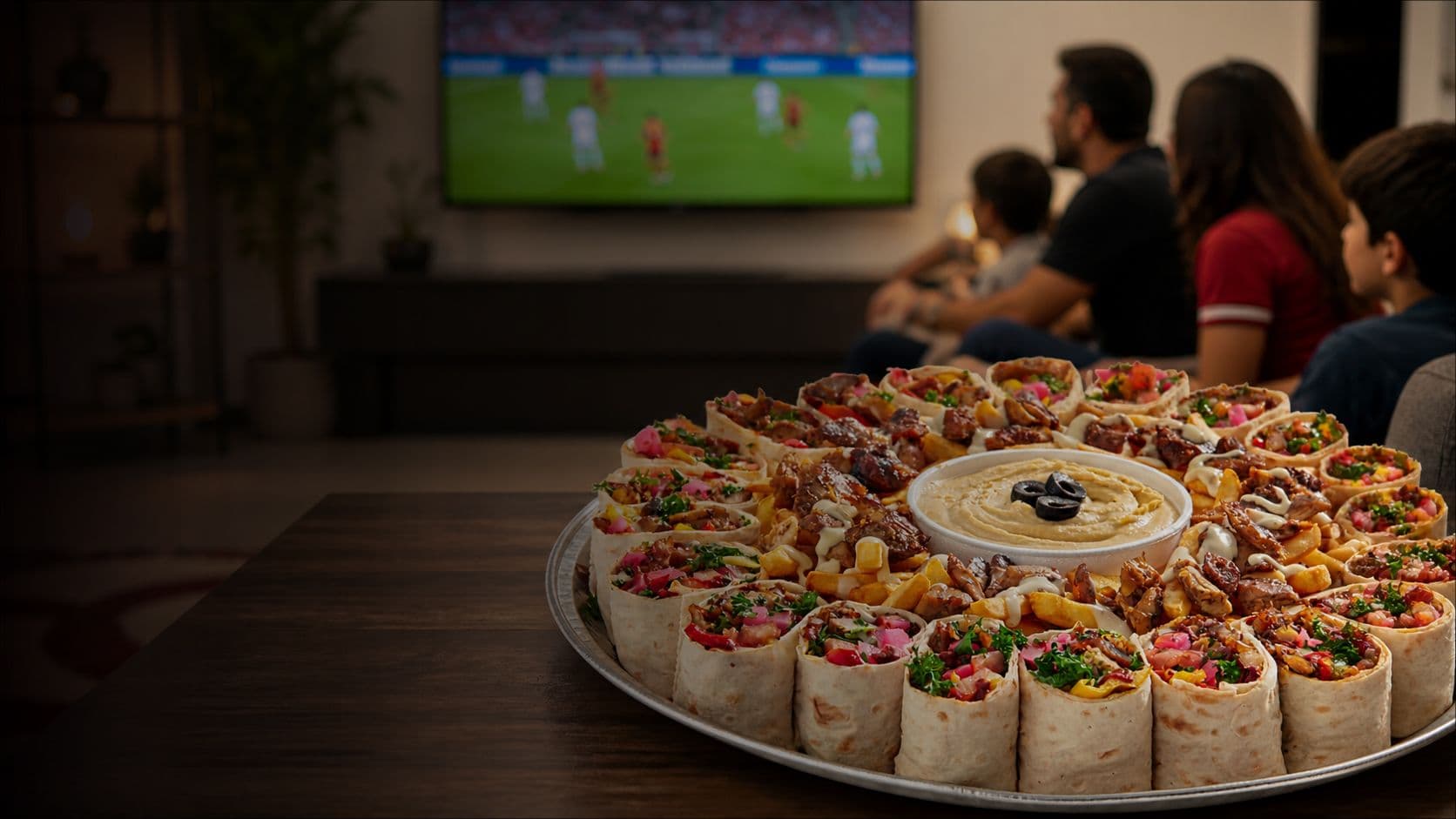 A Lazeez catering spread on a table with family watching tv